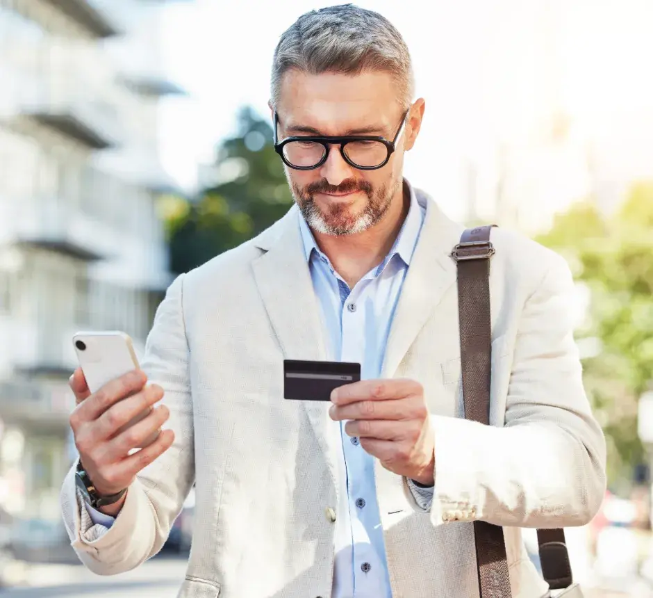 A man holding a credit card while checking his phone on a city street